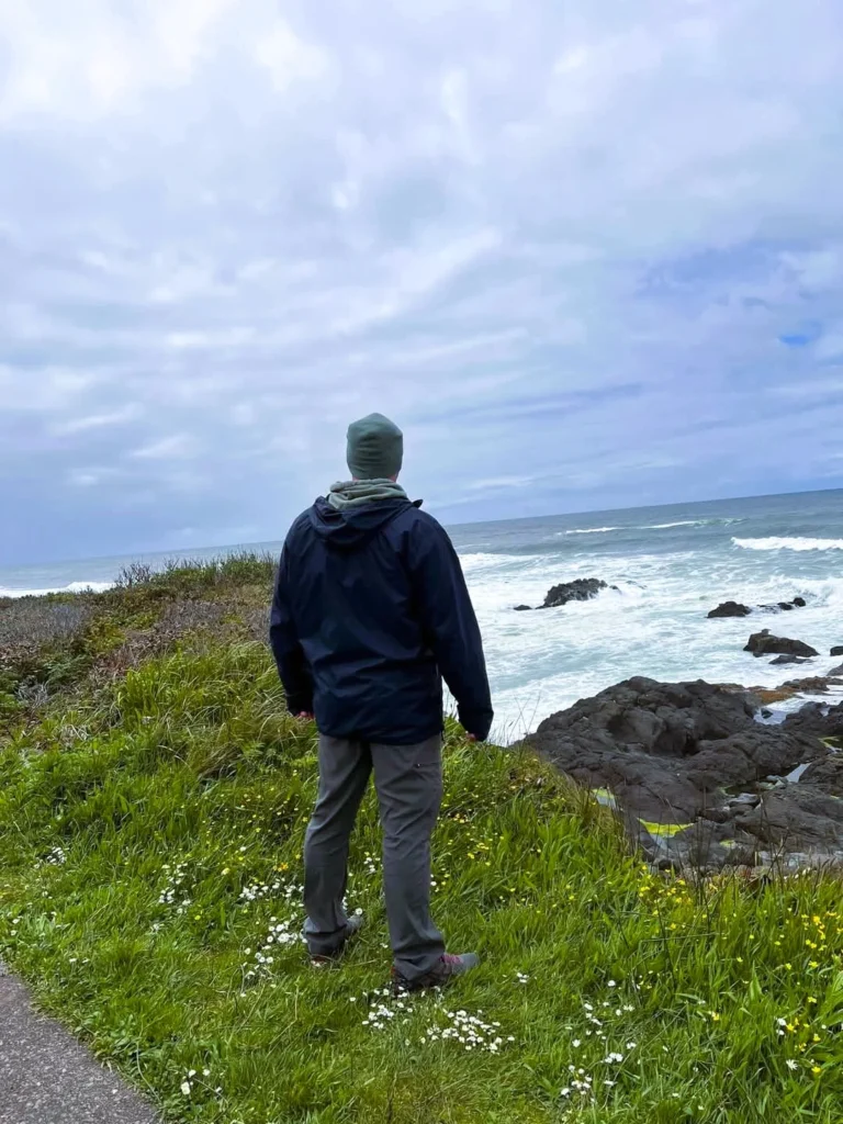Person standing on a rocky overlook on the Oregon Coast with waves crashing below under a cloudy sky.