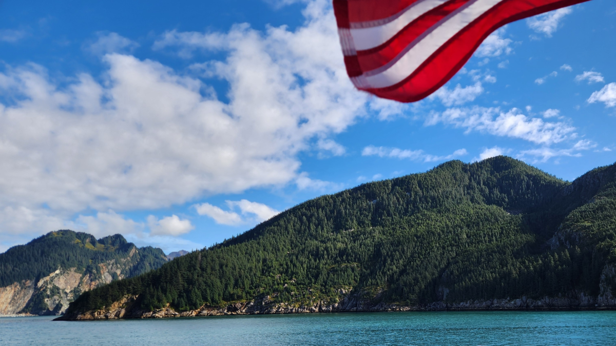 American flag waving on a Major Marine Tours vessel cruising along the forested coastline of Kenai Fjords National Park near Seward, Alaska.