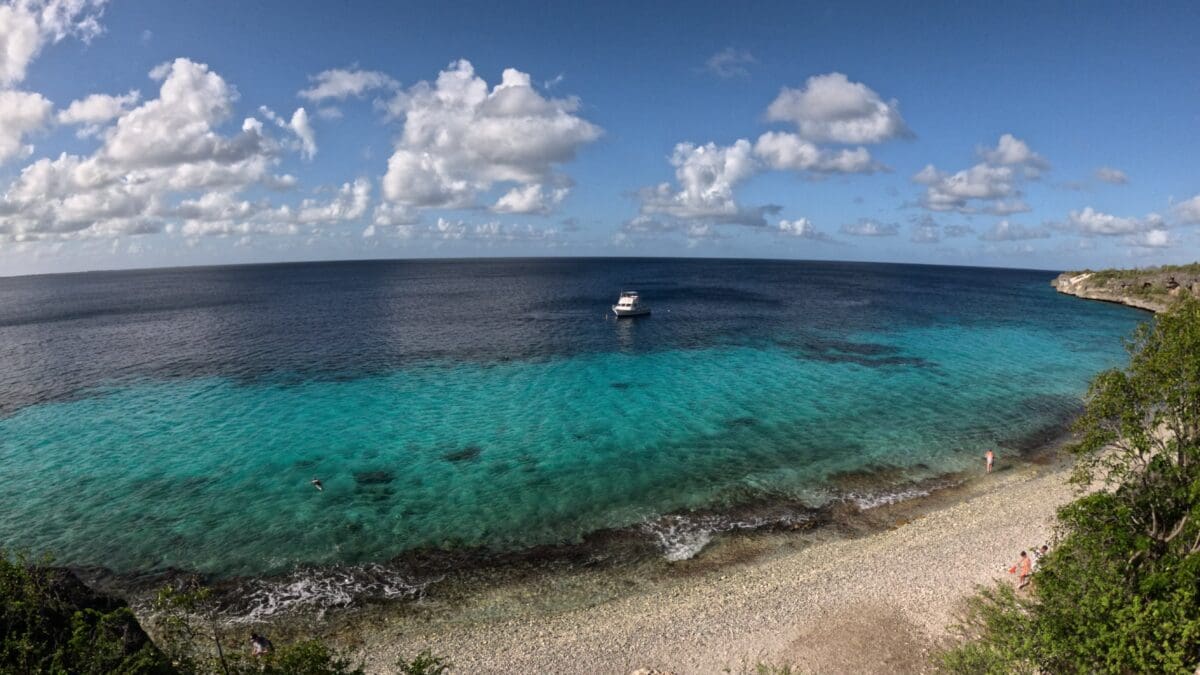 1000 Steps Beach Bonaire from Shore - Dohrn Travels Clear turquoise water at 1000 Steps Beach in Bonaire with a boat offshore and rocky shoreline