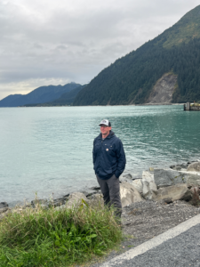 Man in a dark jacket and cap stands on a rocky lakeshore beside a calm turquoise lake, with forested mountains in the background.