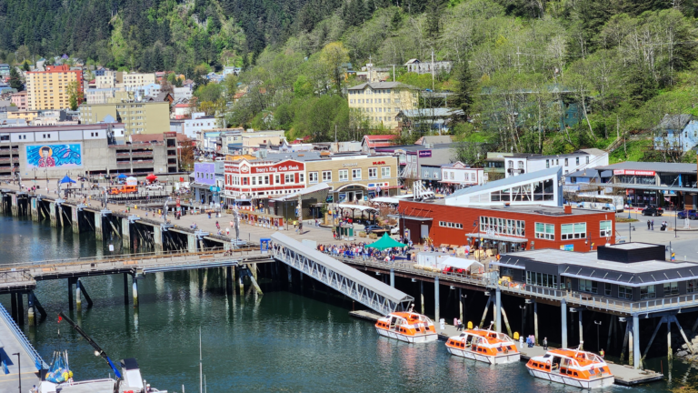 Coastal town along a pier with colorful buildings and orange boats on calm water, backed by green hills and trees in the background.