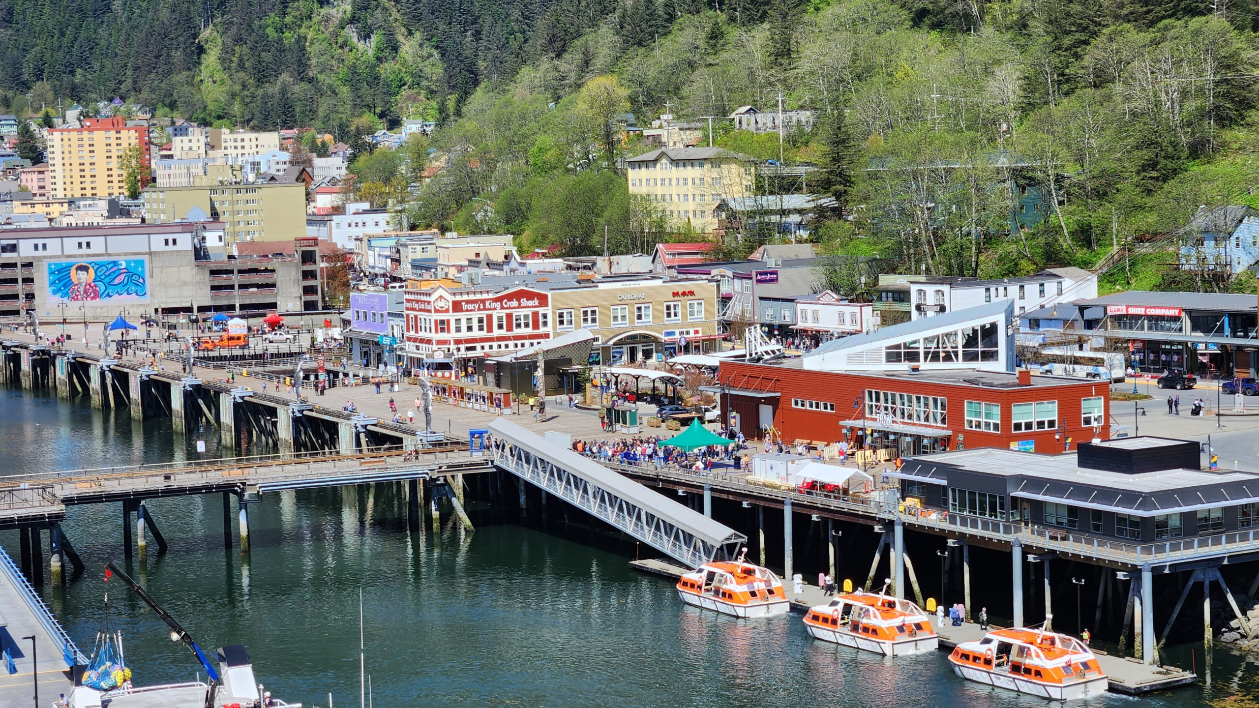 Coastal town along a pier with colorful buildings and orange boats on calm water, backed by green hills and trees in the background.