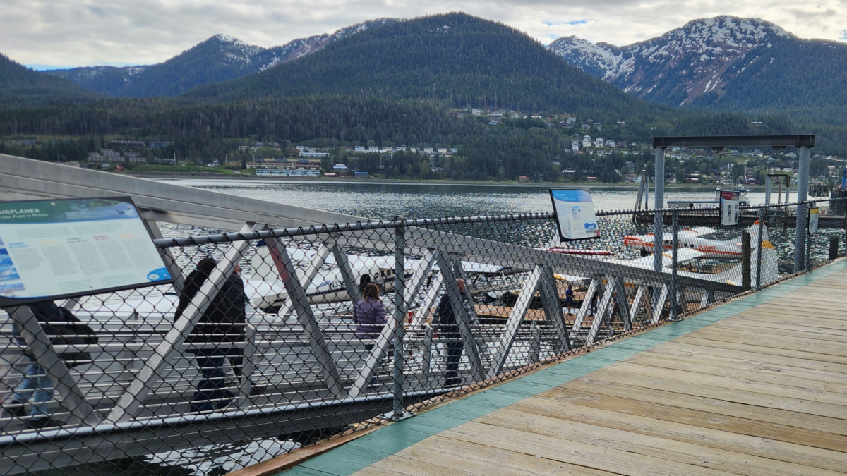 View of a wooden dock with boats moored along a chain-link fence, lake and forested mountains in the background