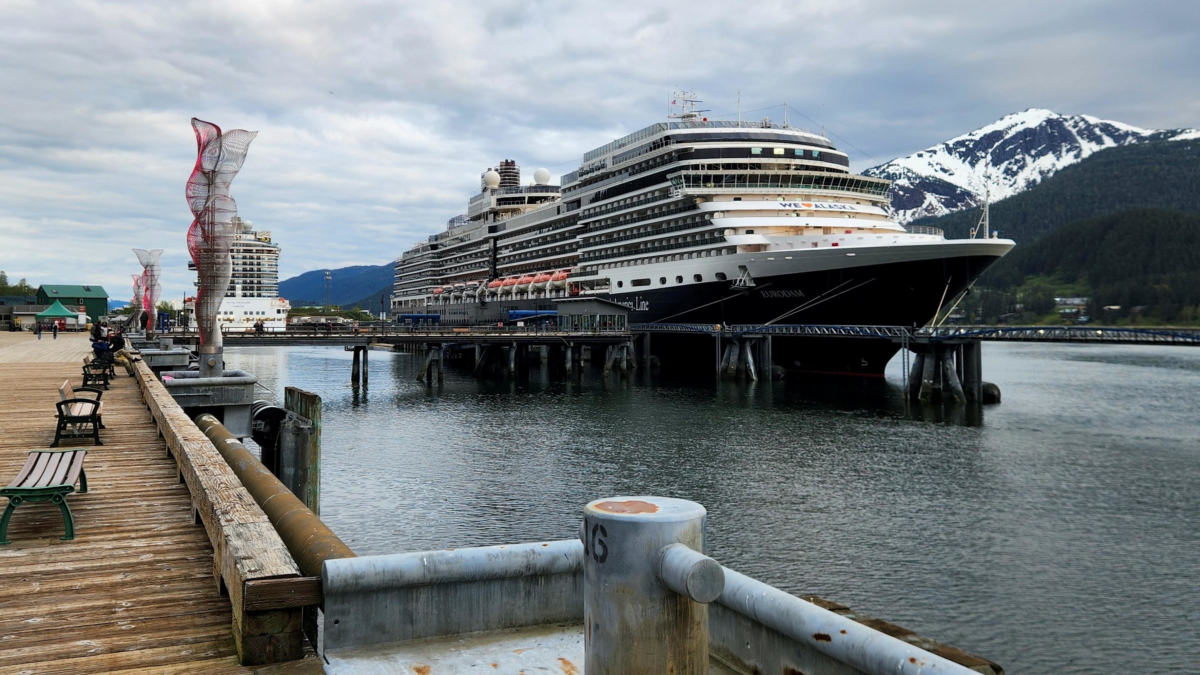 Large cruise ship docked at a pier with a mountainous backdrop and calm water, benches along the wooden boardwalk in the foreground.