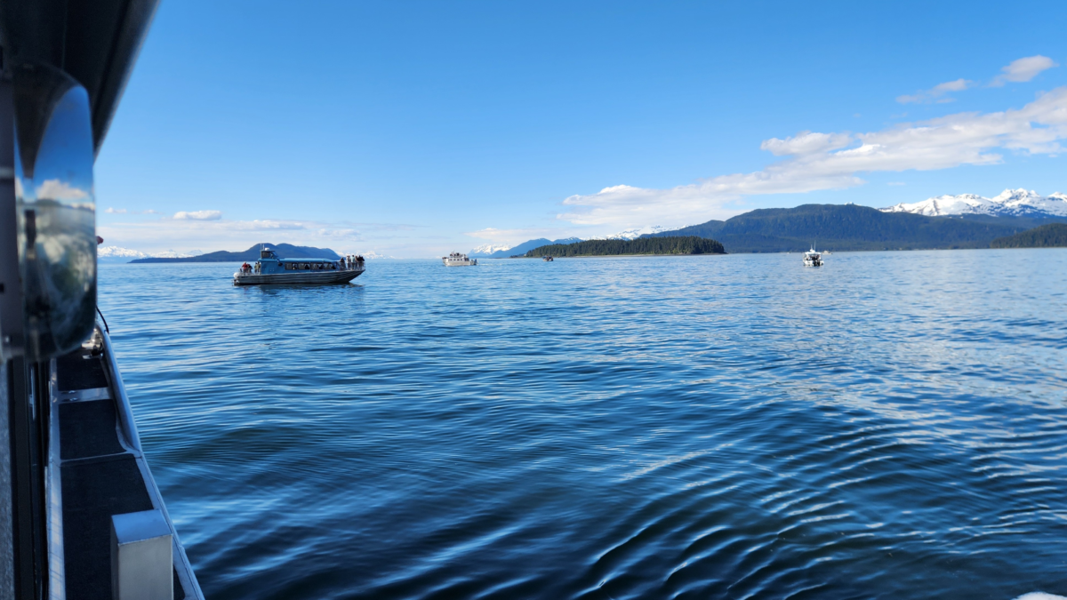 Boat on a calm blue lake with distant snow-capped mountains and a clear sky, photographed from another boat's side.