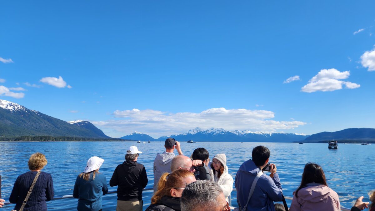 People on a boat enjoying a lake view with snow-capped mountains in the distance and a clear blue sky above.