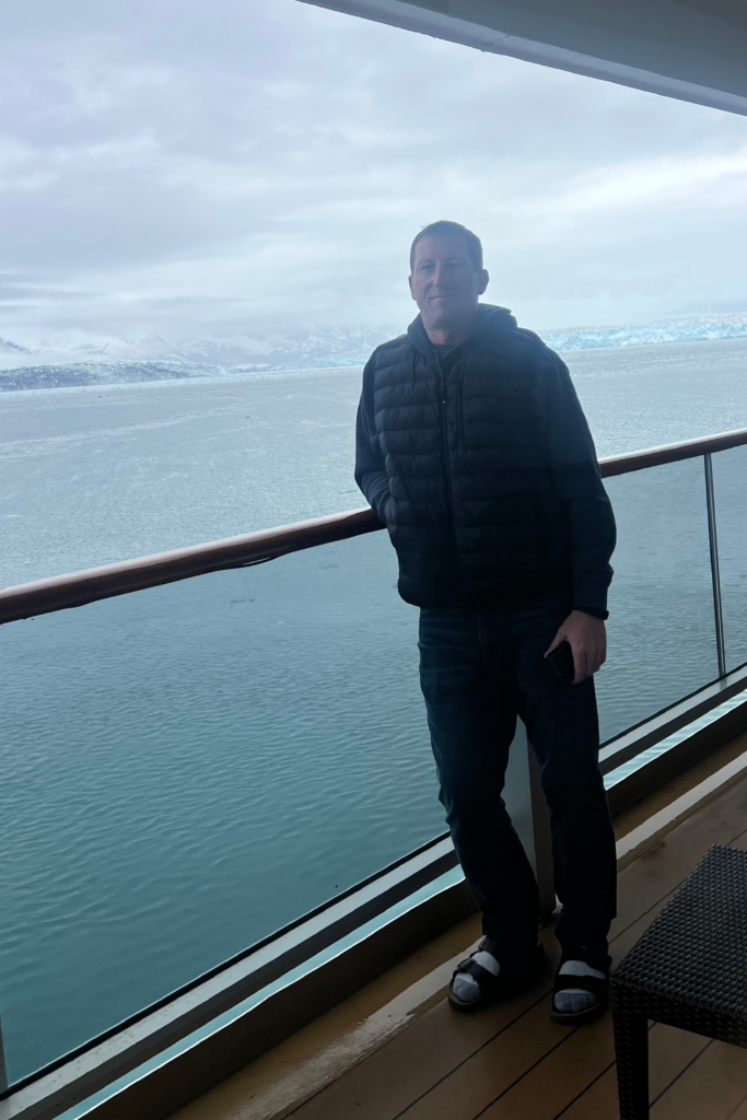 Layering Near Hubbard Glacier on an Alaska Cruise | Dohrn Travels Man wearing a puffer vest and hoodie on a cruise ship balcony near Hubbard Glacier in Alaska.