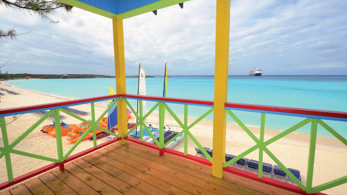 View from a colorful beach villa balcony at Half Moon Cay, overlooking the white-sand beach, turquoise water, and a cruise ship offshore.